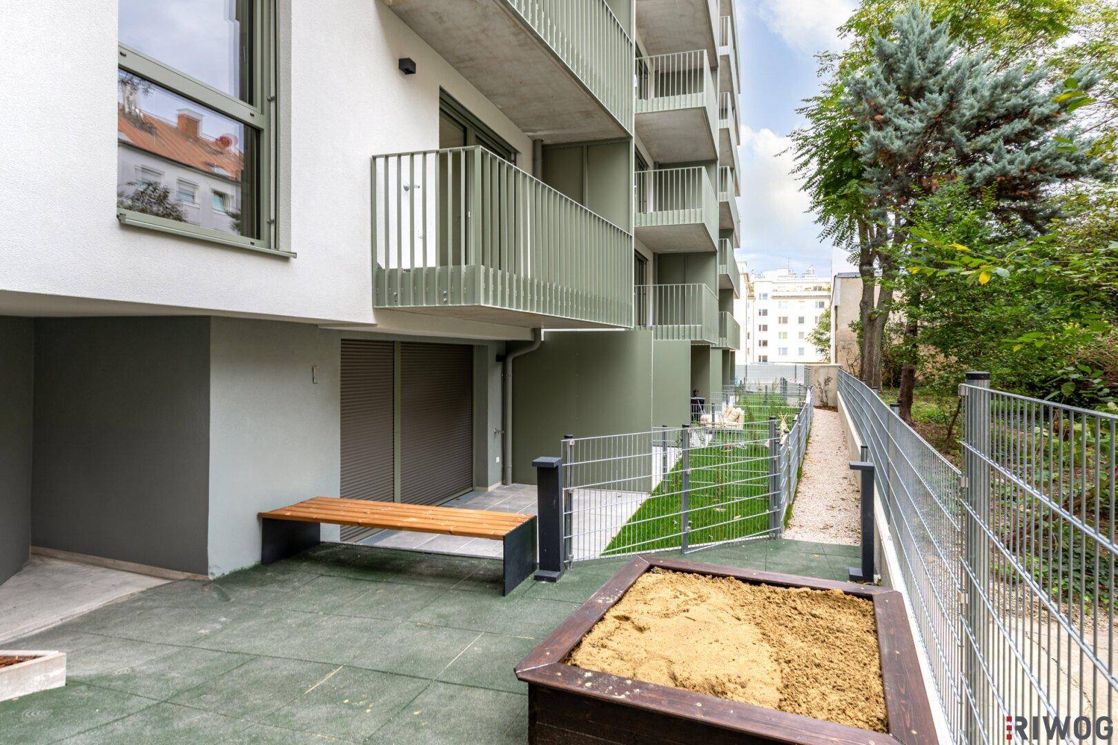 Außenbereich mit Sandkasten und Sitzbank, umgeben von Grünflächen und einem modernen Gebäude.