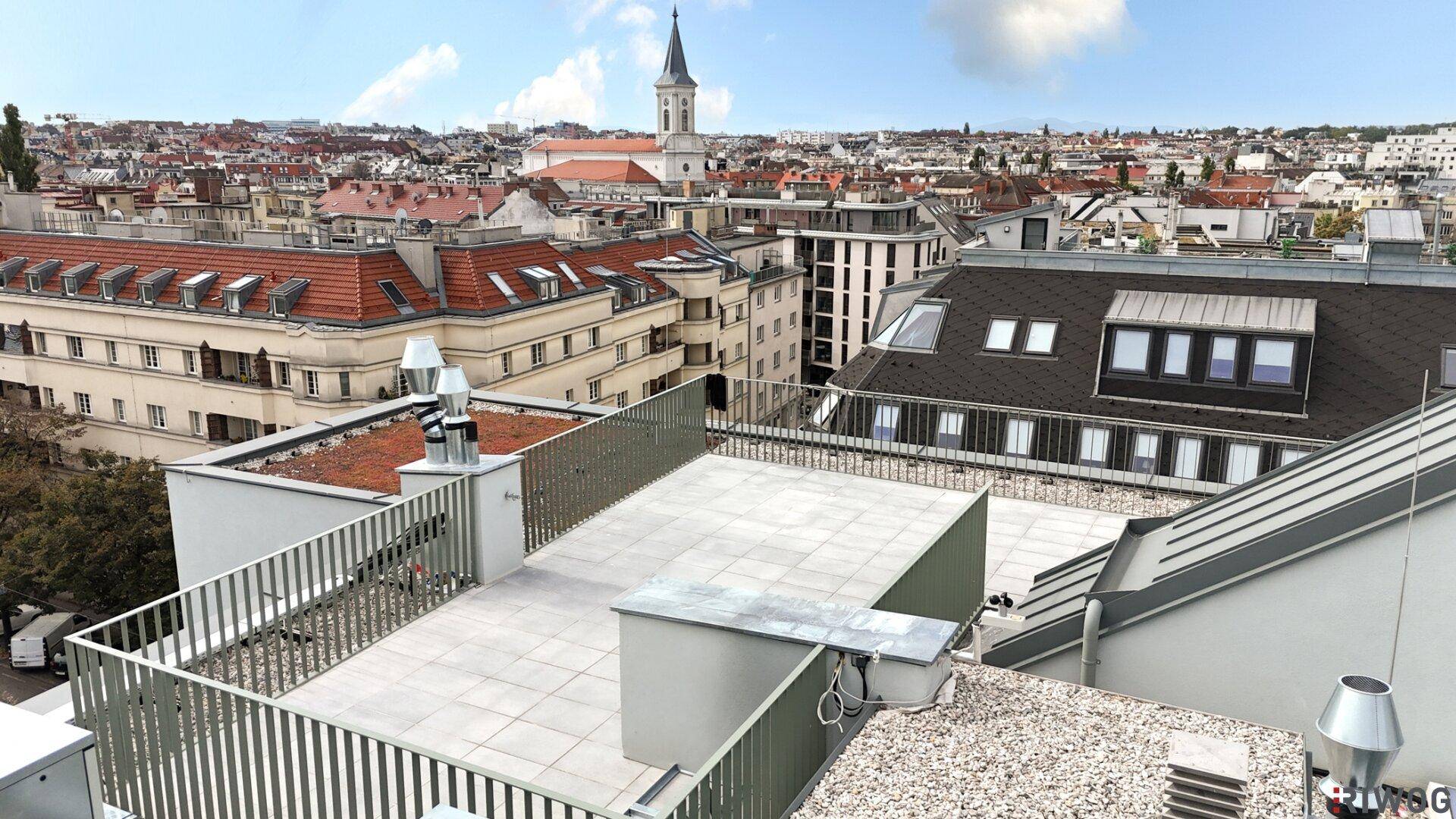 Dachterrasse mit Blick auf die Stadt, umgeben von anderen Dächern und einem Kirchturm.