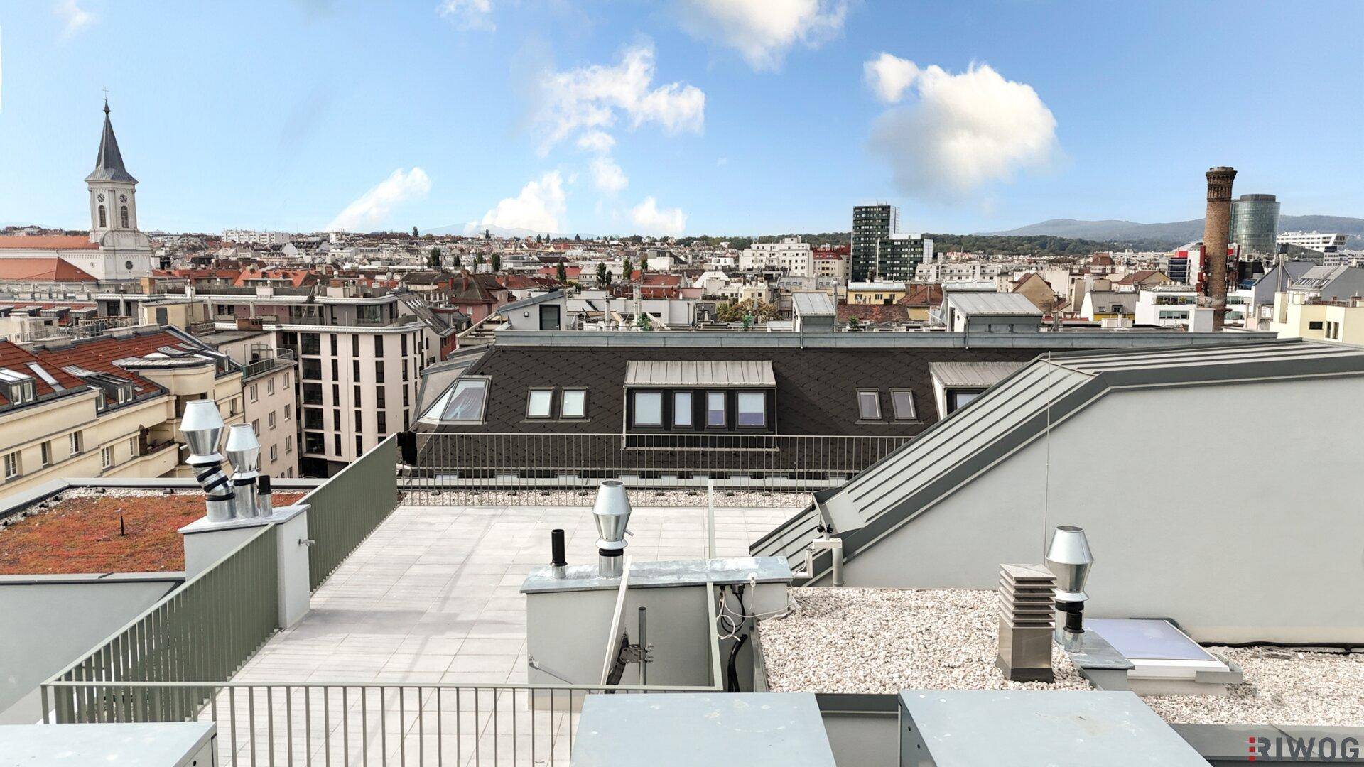 Dachterrasse mit weitem Blick über die Stadt, umgeben von Dächern und einem Kirchturm.
