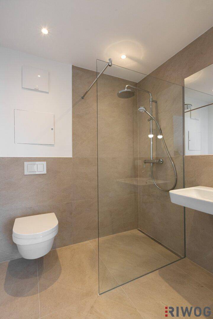 Zeitgemäßes Badezimmer mit großer Glasdusche, Wand-WC und beigen Wand- und Bodenfliesen.