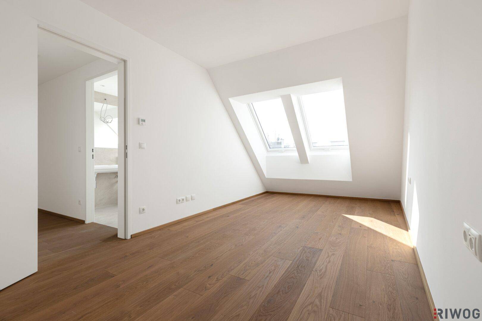 Heller Dachgeschossraum mit Holzboden und Dachfenstern, offen zu einem Nebenraum.