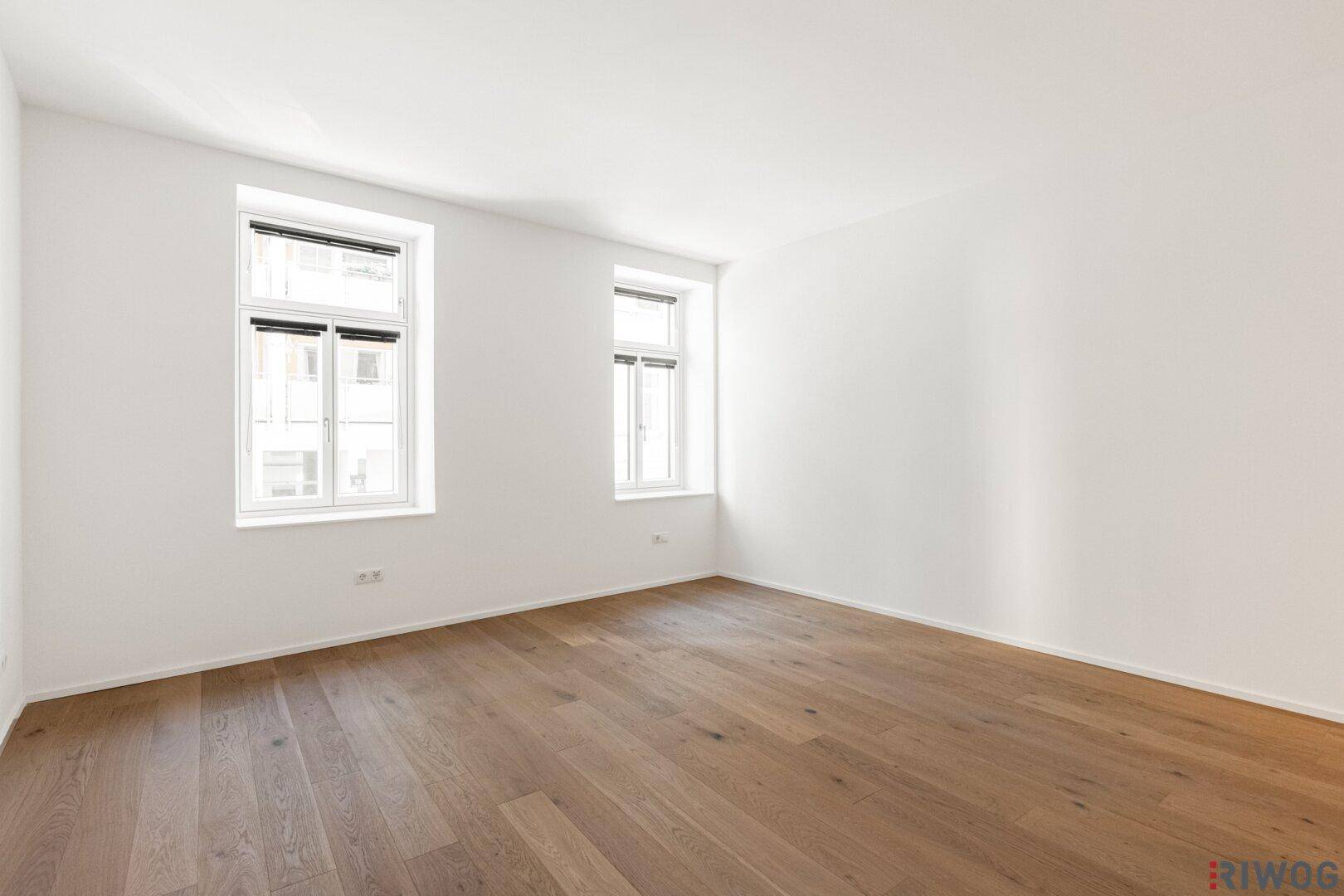 Heller, leerer Raum mit Holzboden und zwei großen Fenstern, bereit zur individuellen Gestaltung.