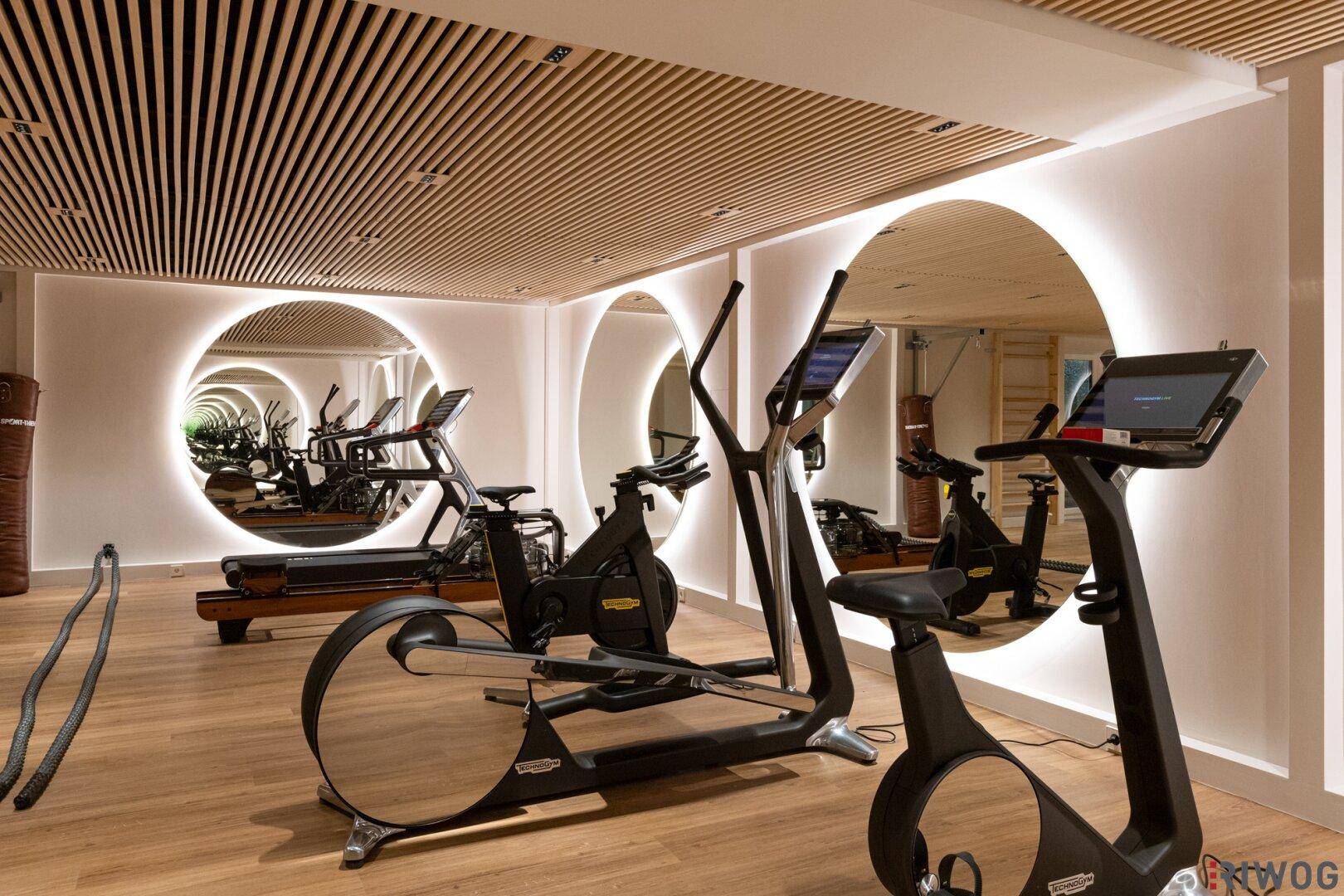 Fitnessraum mit Laufband und Rudergerät, umgeben von modernen Spiegelflächen und Holzdecken.