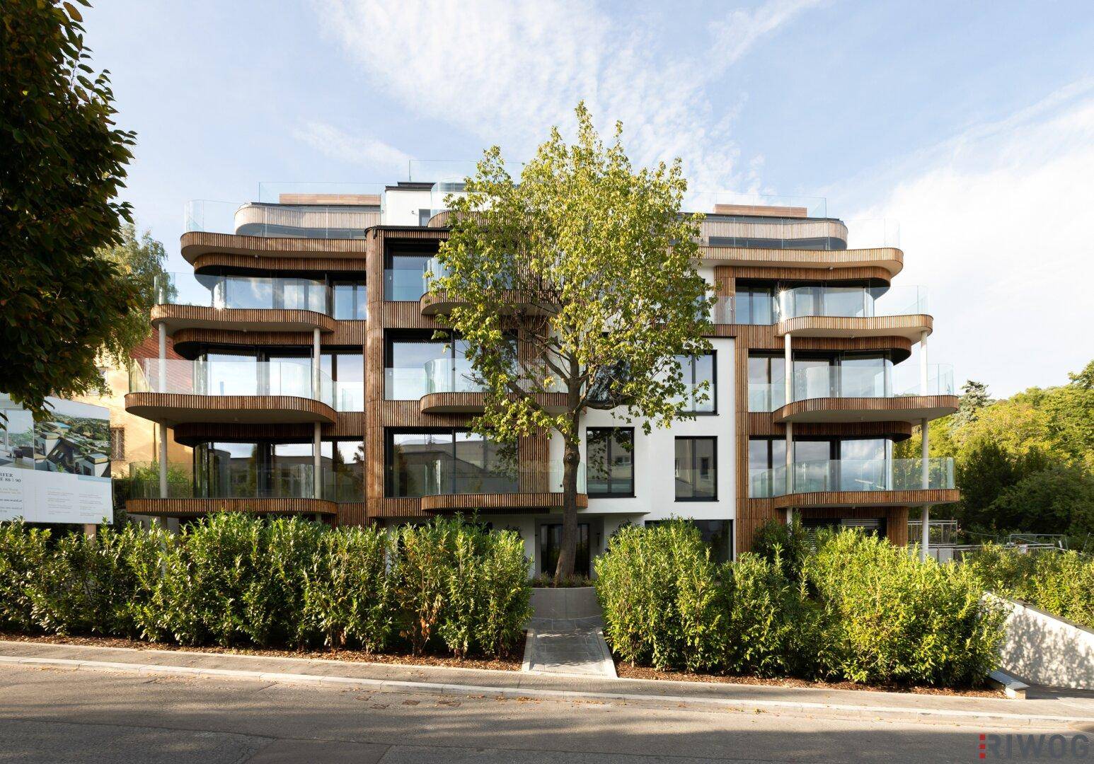 Modernes Wohngebäude mit Holzfassade, Balkonen und Tiefgaragenzufahrt, umgeben von Bäumen.