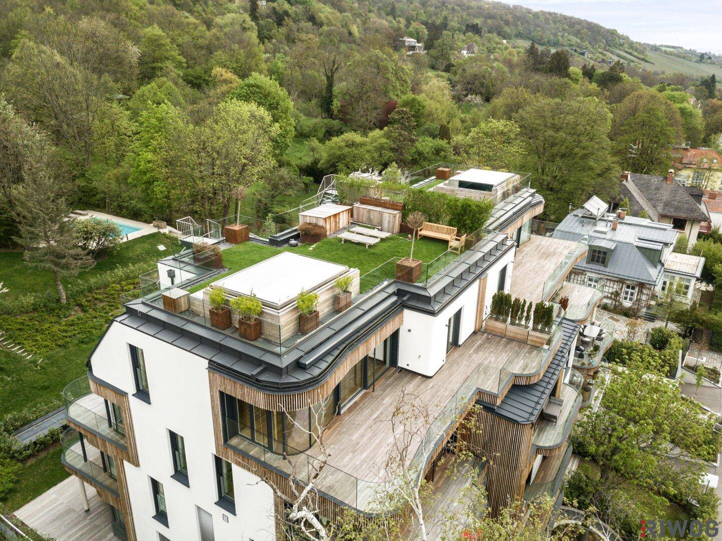 Luftaufnahme eines modernen Wohnkomplexes mit grünen Dächern und Terrassen, umgeben von Bäumen und Stadtblick.