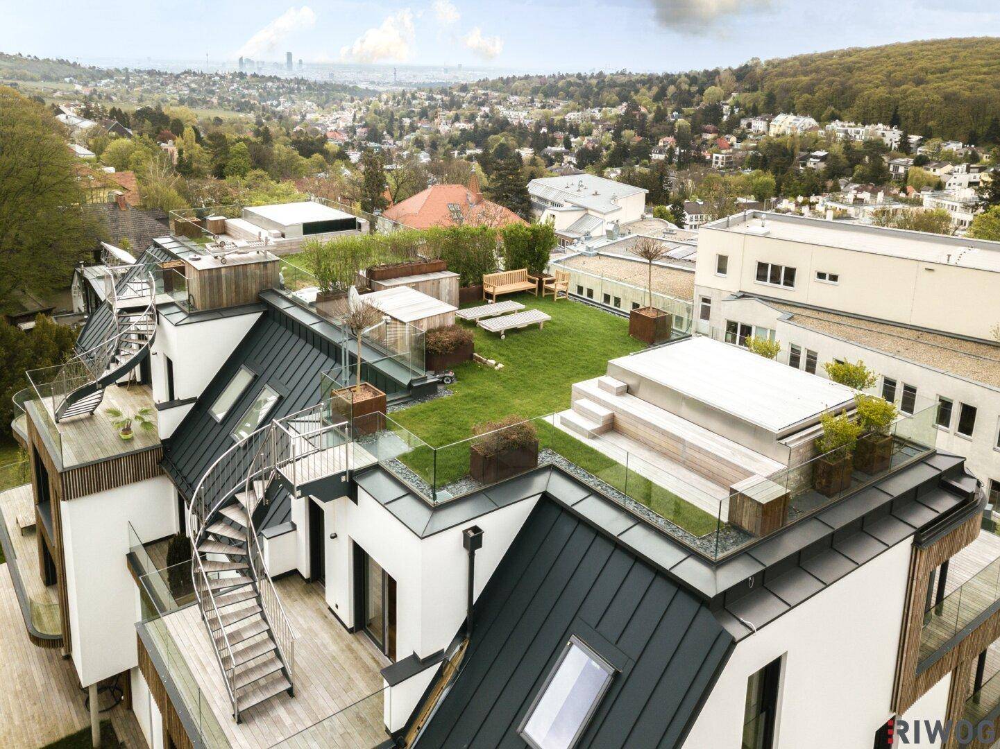 Vogelperspektive eines modernen Gebäudes mit Holzfassade und großzügigen Terrassen, umgeben von viel Grün.