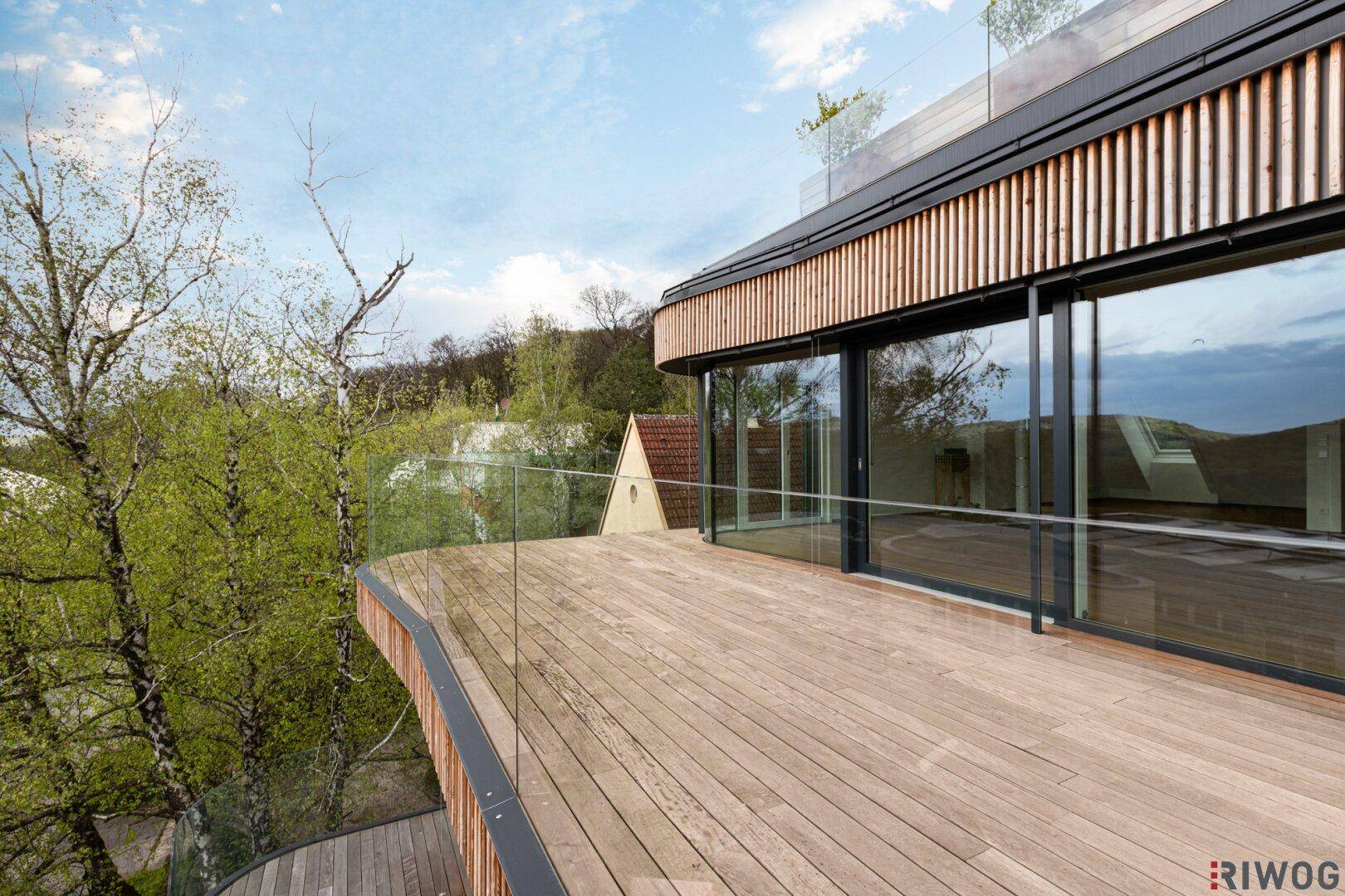 Weitläufige Holzterrasse mit elegantem Glasgeländer und umgebender Natur, bietet viel Platz im Freien.