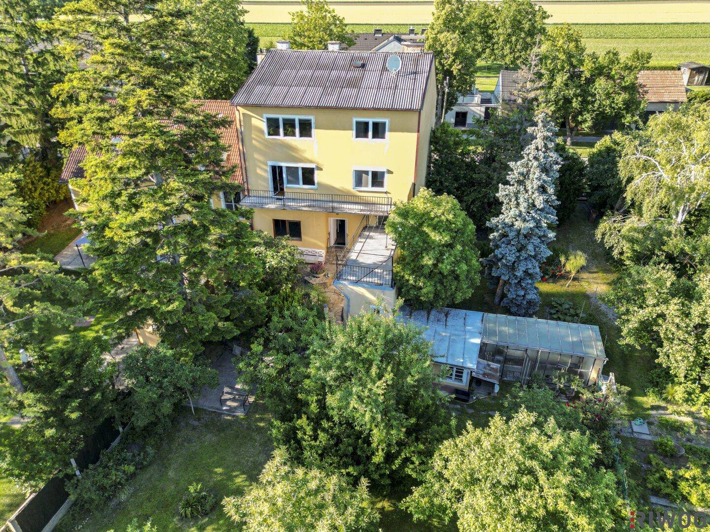 Rückseite eines gelben Hauses mit mehreren Terrassen und einem Gewächshaus im Garten.