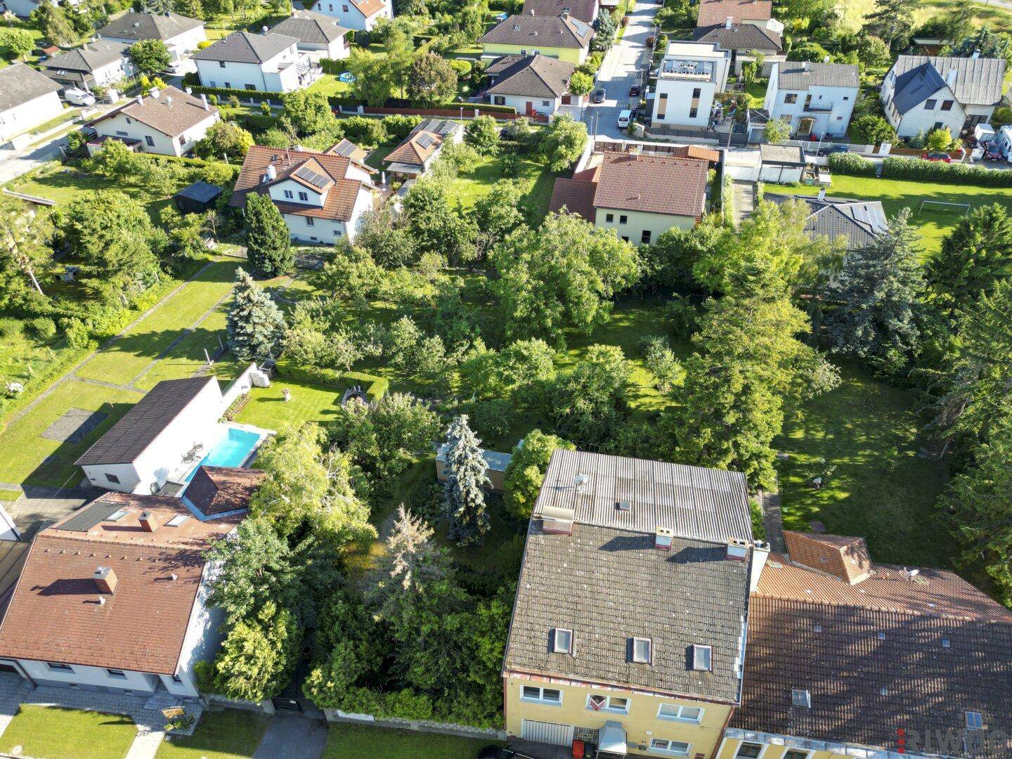 Luftaufnahme einer Wohnsiedlung mit einem gelben Haus im Vordergrund und umliegenden Gärten.