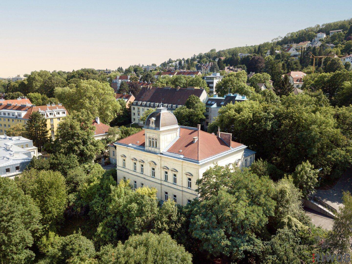 Weitläufige Luftaufnahme der historischen Villa in einer grünen, hügeligen Stadtlandschaft.