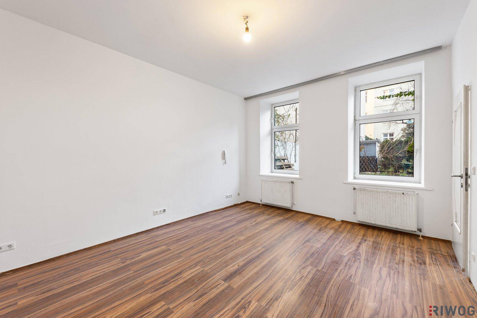 Geräumiges Wohnzimmer mit Holzboden und zwei großen Fenstern, die viel Tageslicht hereinlassen.