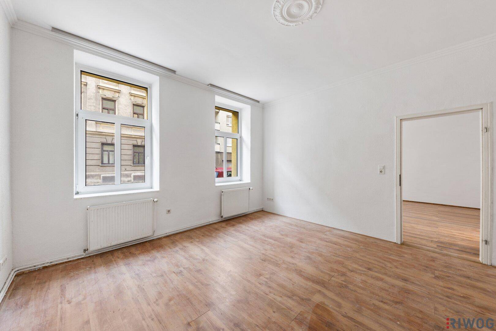 Heller Wohnbereich mit Holzboden und Stuckdecke, der Zugang zu einem weiteren Raum bietet.