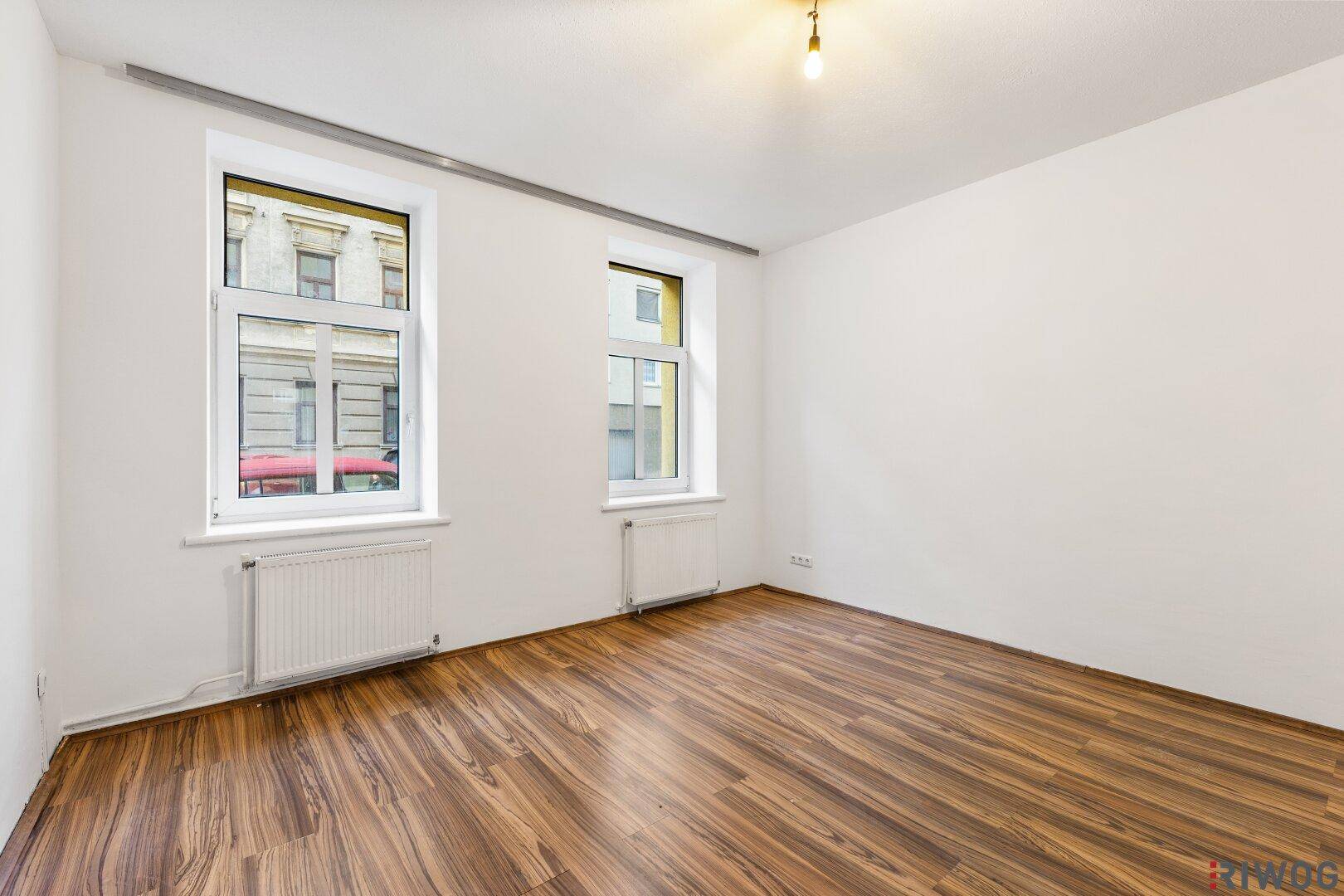 Helles Wohnzimmer mit Holzboden und zwei Fenstern, die viel Tageslicht hereinlassen.