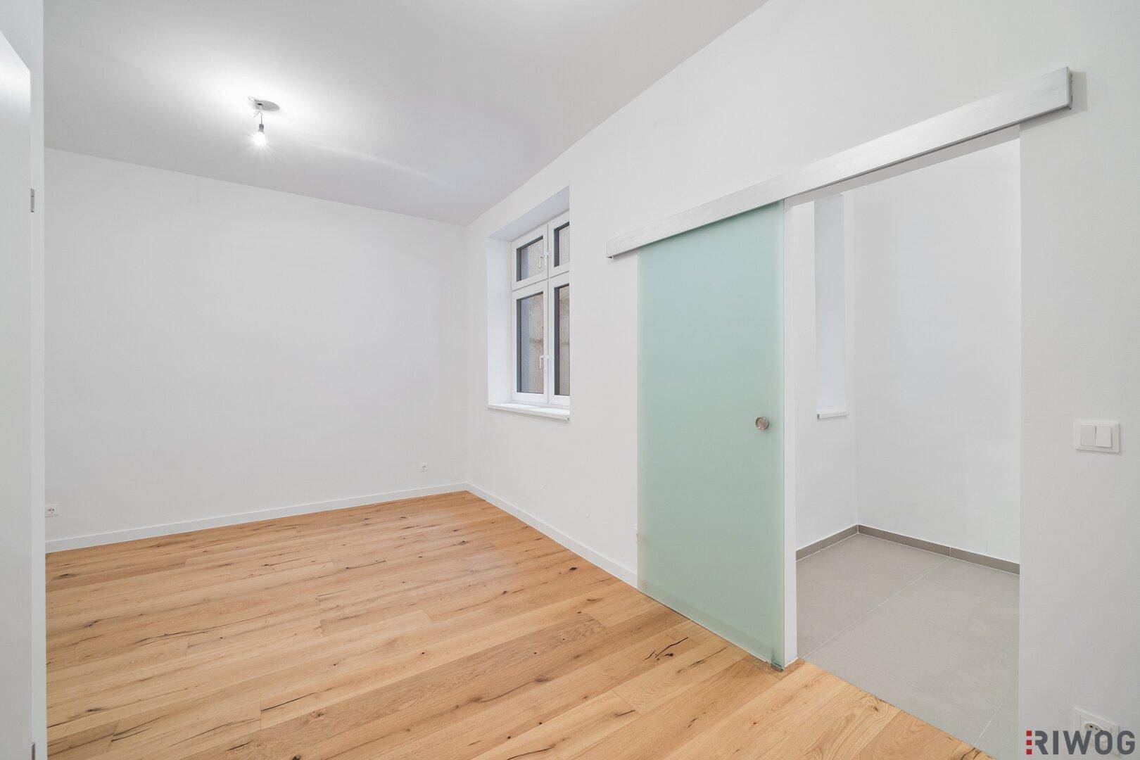 Heller Raum mit Holzboden und einer modernen Glasschiebetür, die zu einem weiteren Bereich führt.