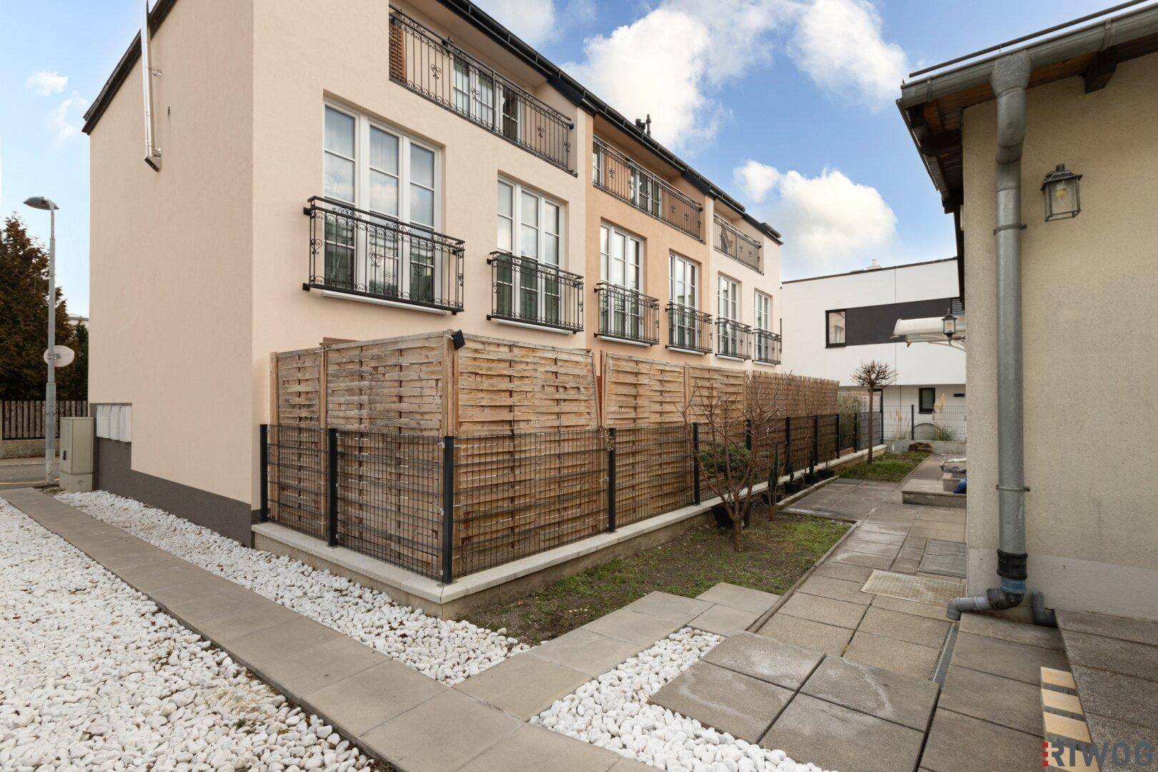 Seitenansicht des Gebäudes mit einem kleinen Gartenstreifen, Holzzaun und gepflastertem Weg.