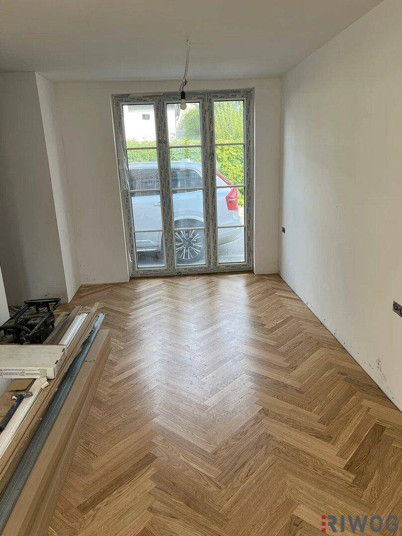 Heller Raum mit Fischgrätparkett und großen Fenstertüren, die viel Tageslicht hereinlassen, noch unmöbliert.