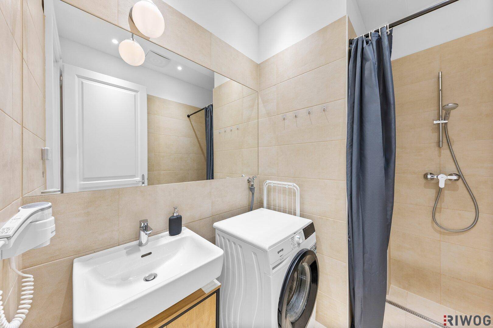 Modernes Badezimmer mit beigefarbenen Fliesen, Dusche, Waschbecken und integrierter Waschmaschine.
