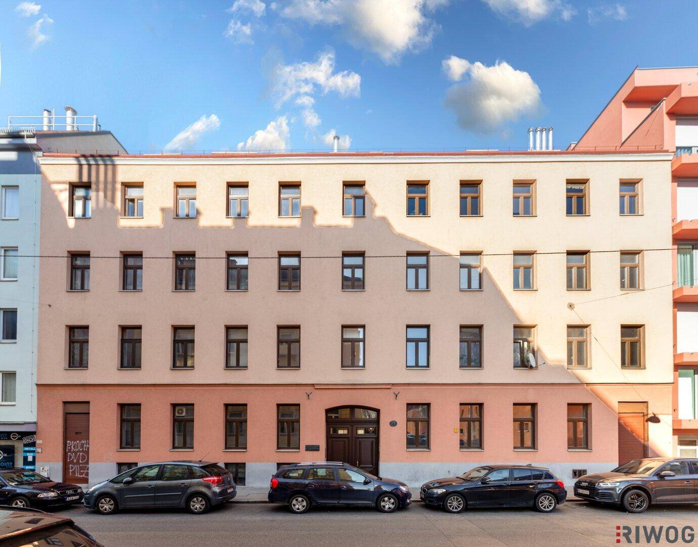 Historische Fassade eines Mehrfamilienhauses mit vielen Fenstern und parkenden Autos auf der Straße.