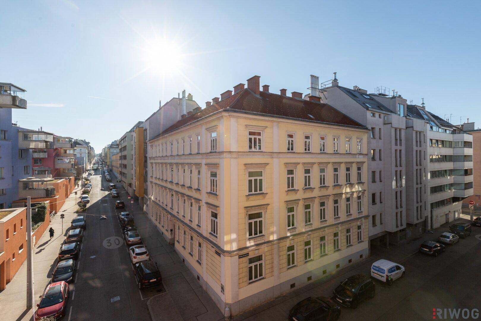 Historisches Eckgebäude mit gelber Fassade und vielen Fenstern an einer sonnigen Stadtstraße.