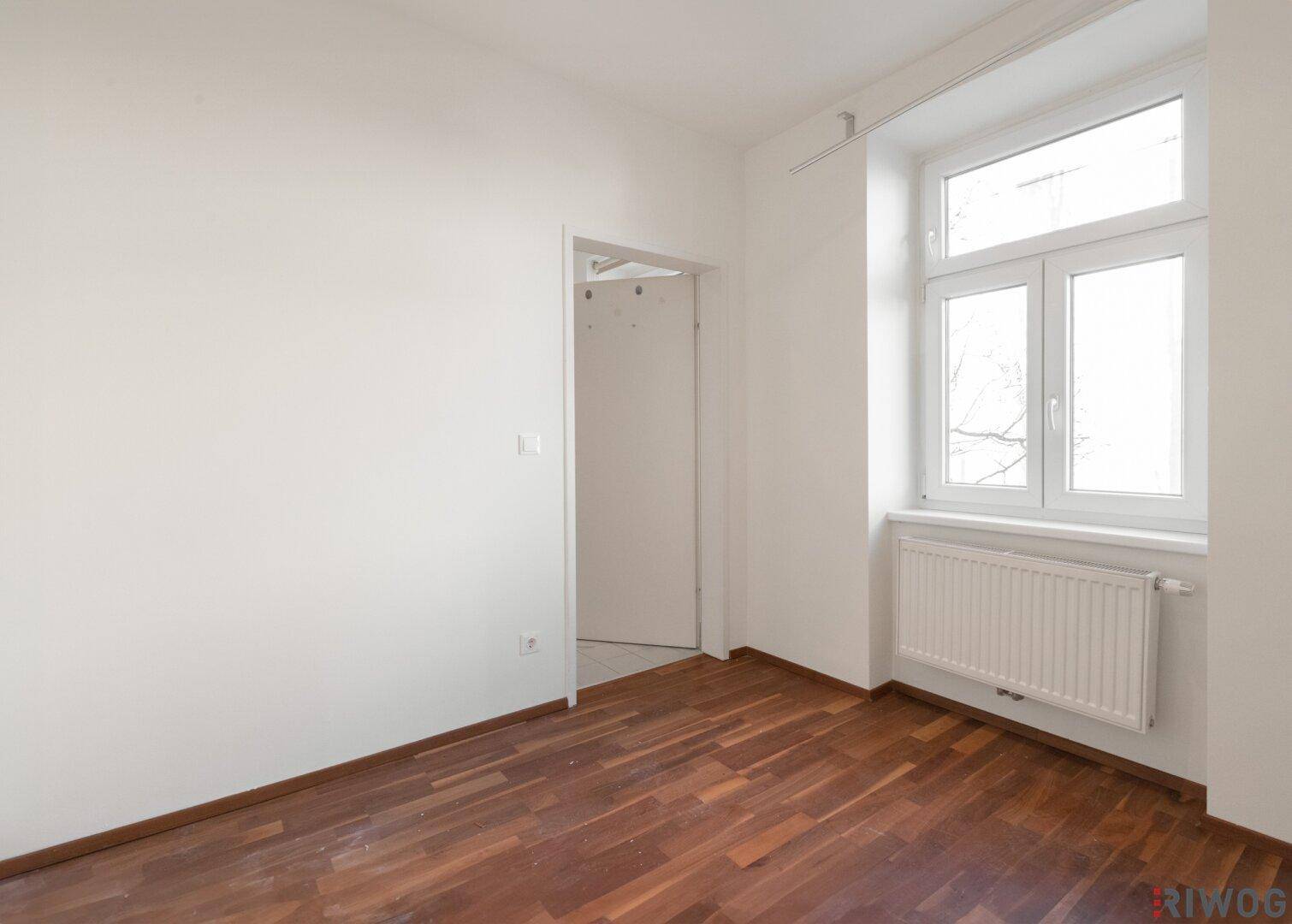 Helles Schlafzimmer mit Parkettboden und einem Fenster, das viel Tageslicht hereinlässt.