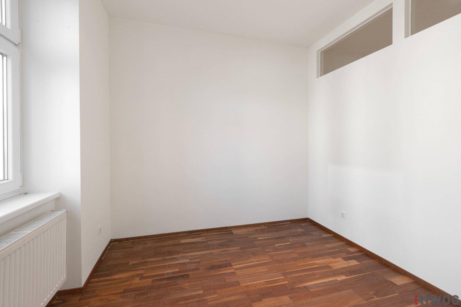 Zimmer mit Holzboden, weiß gestrichenen Wänden und einem hohen Fenster für zusätzliches Licht.
