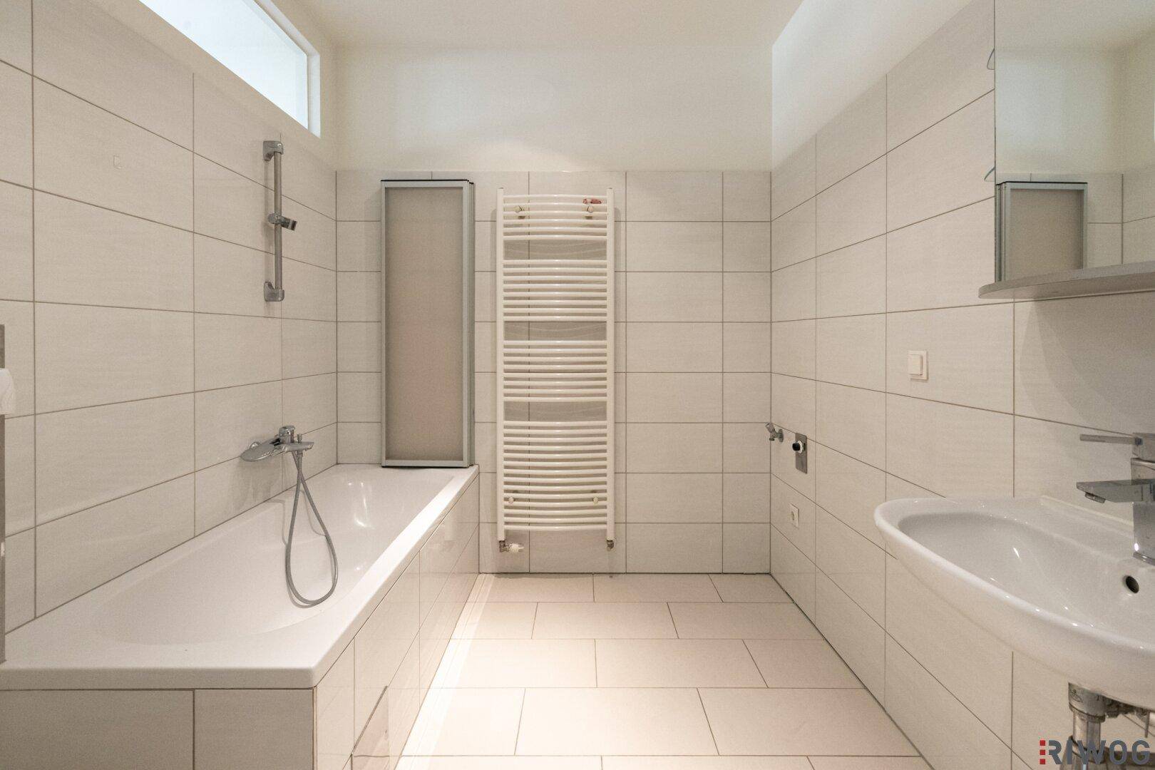 Modernes Badezimmer mit weißen Fliesen, Badewanne, Waschbecken und Handtuchheizkörper.