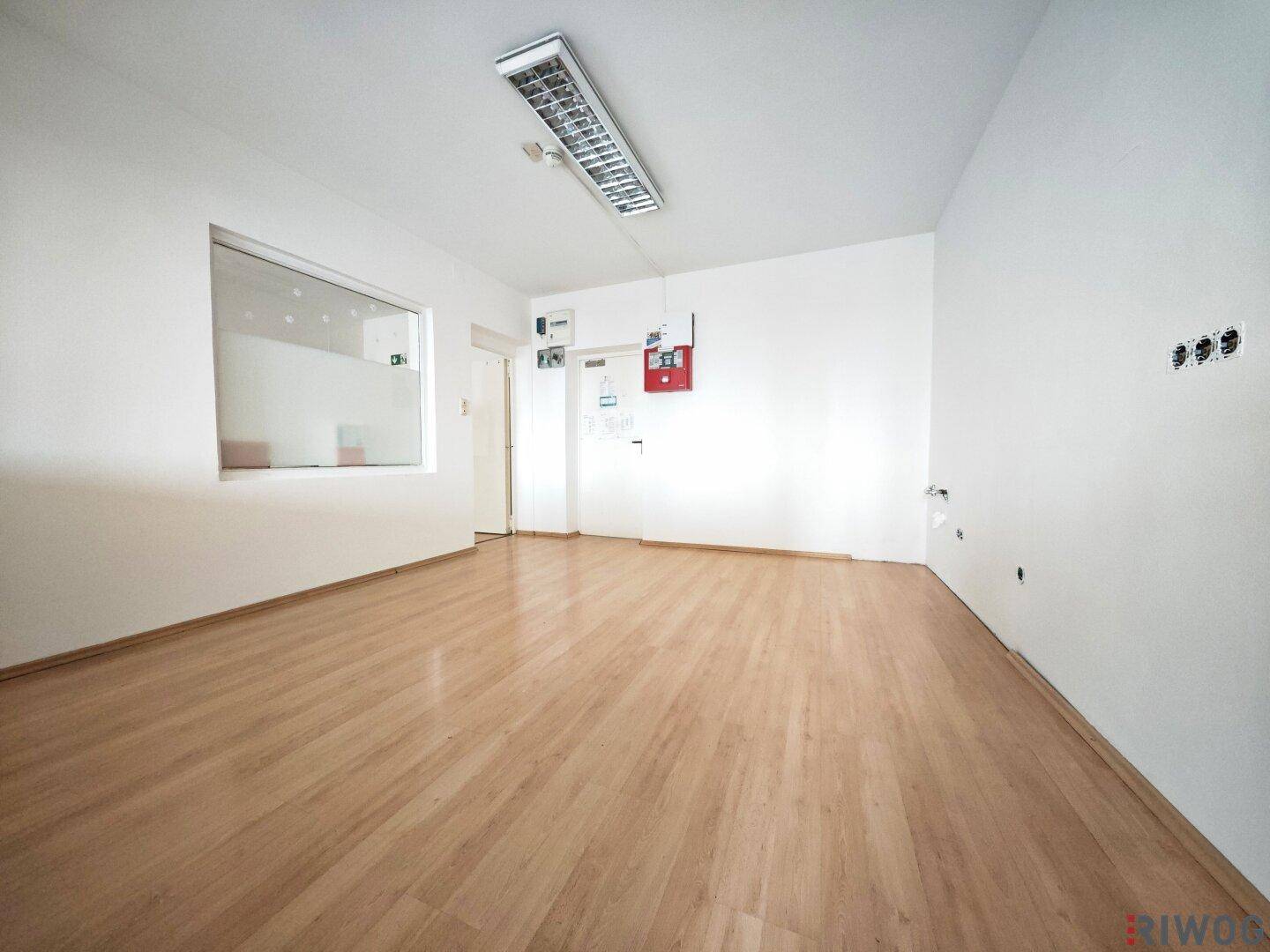 Heller Büroraum mit Laminatboden, weißen Wänden und einem internen Fenster, das Licht hereinlässt.