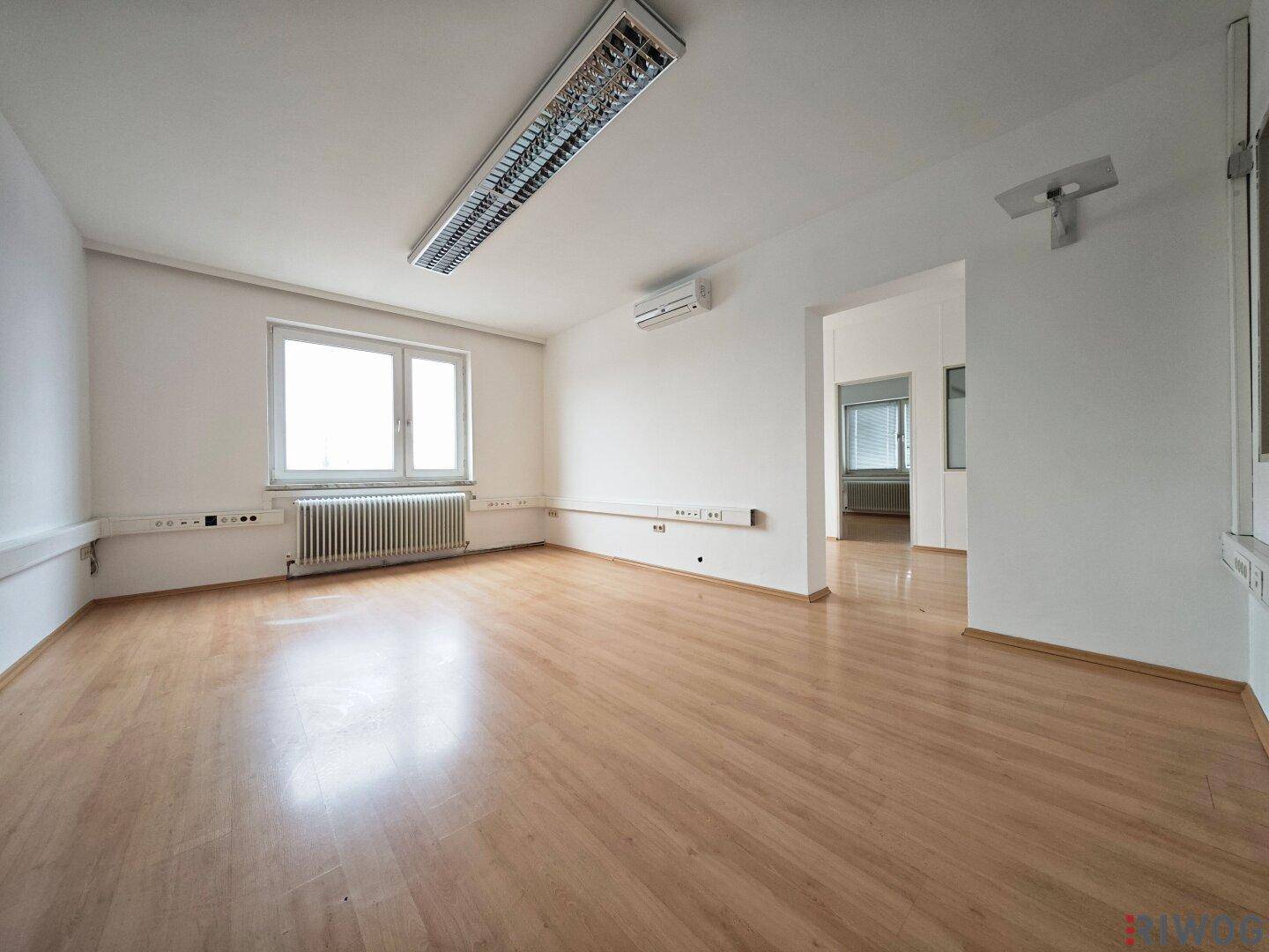 Leerer Büroraum mit Laminatboden, weißen Wänden, Fenster und einer Klimaanlage für angenehmes Klima.