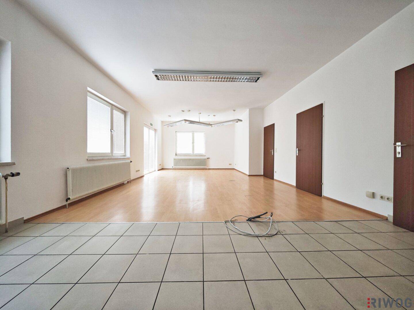 Büroraum mit Übergang von Fliesen- zu Laminatboden, weißen Wänden und Fenstern für natürliche Belichtung.