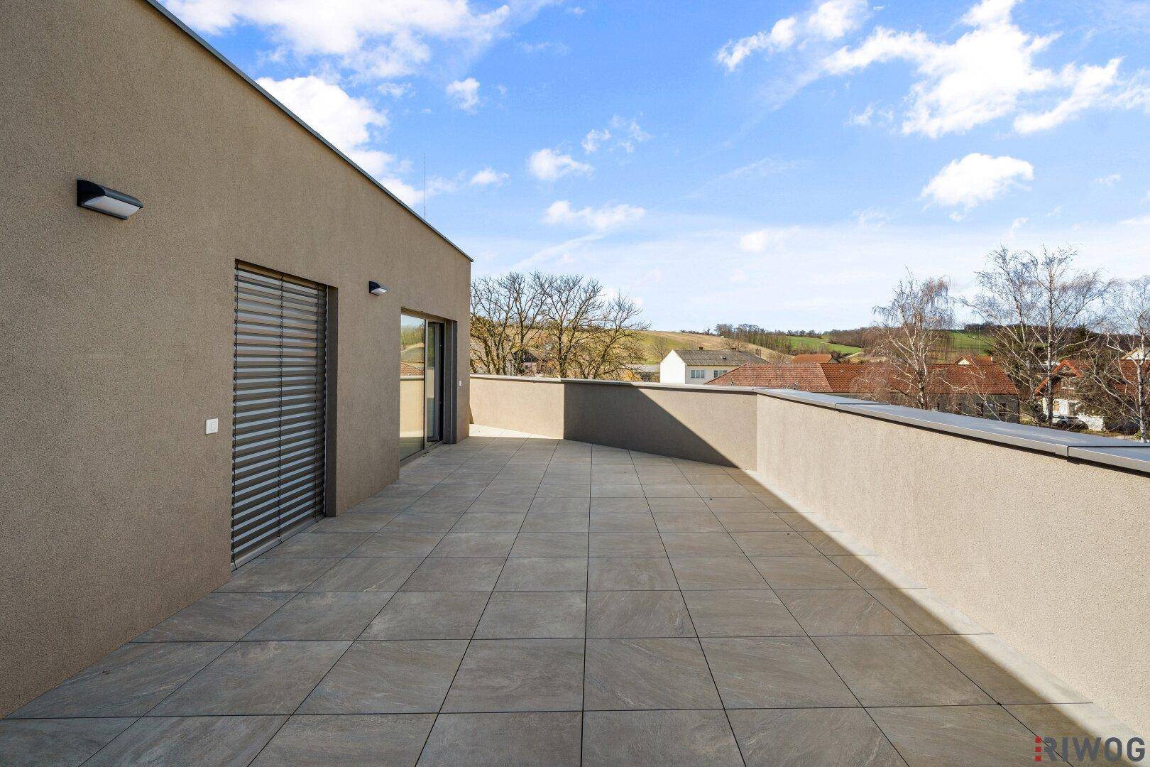 Große, geflieste Terrasse mit Blick auf die umliegende Landschaft und Rollläden an den Fenstern.