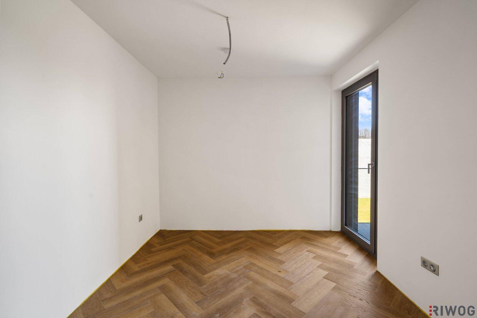 Helles Zimmer mit Fischgrätparkett und hoher Tür, die nach außen führt.