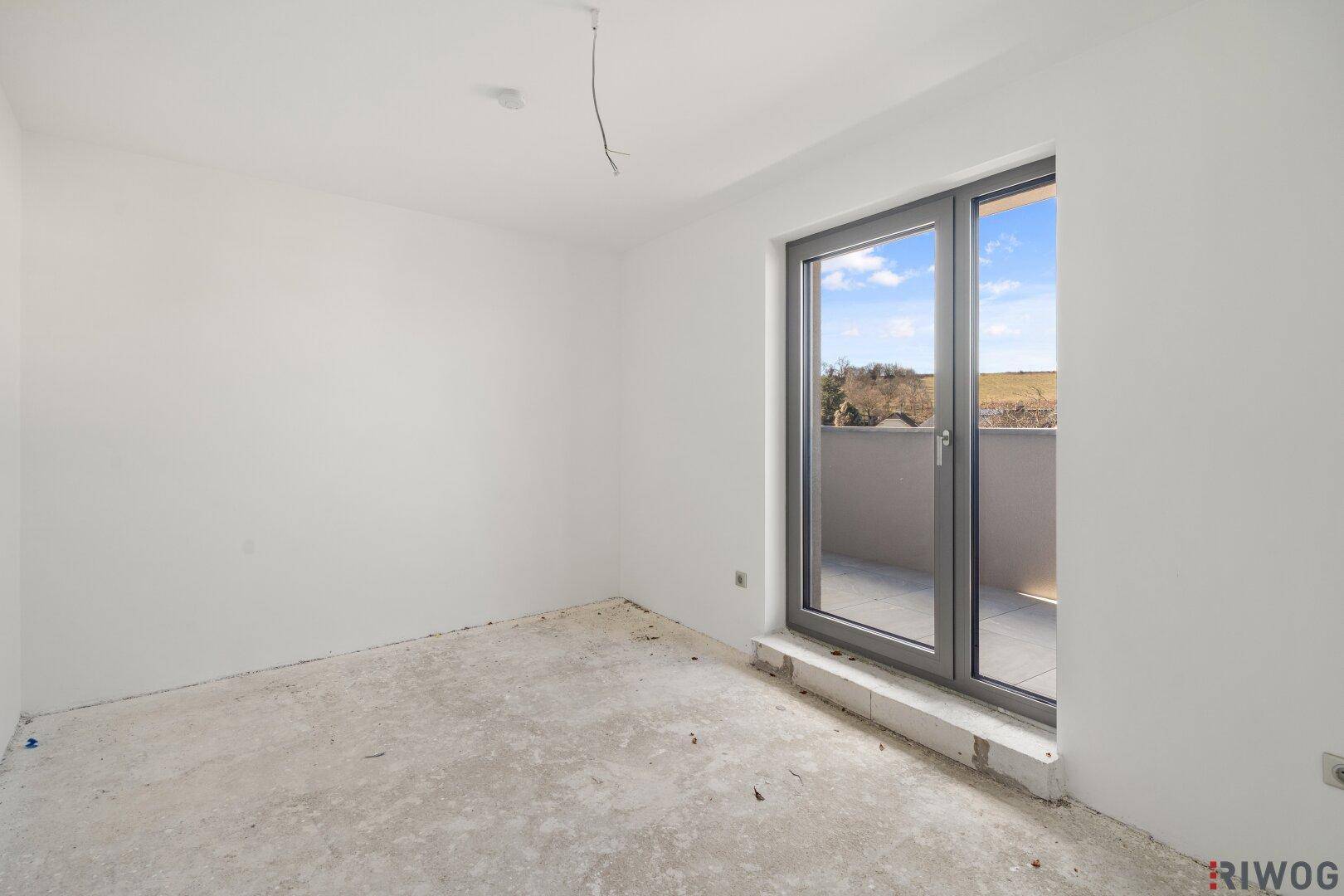 Heller Raum im Rohbau mit Betonboden und Fenster mit Blick ins Grüne.