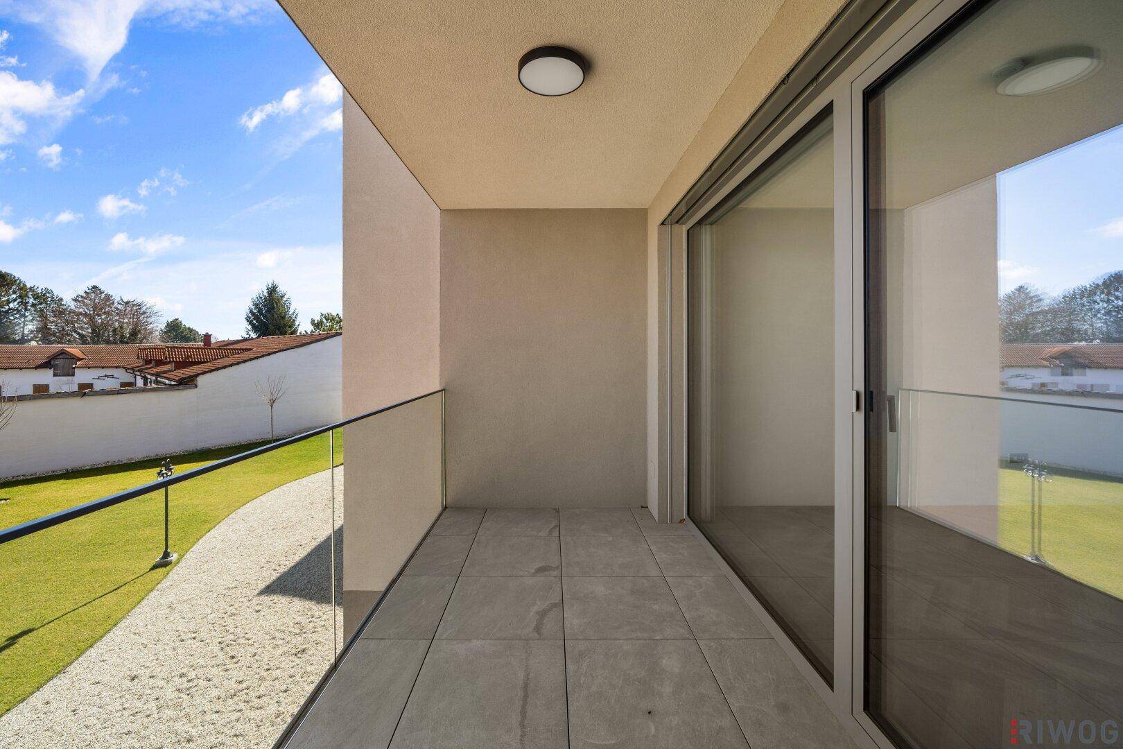 Überdachter Balkon mit Fliesenboden und Glasgeländer, bietet einen Blick auf die gepflegte Grünfläche.