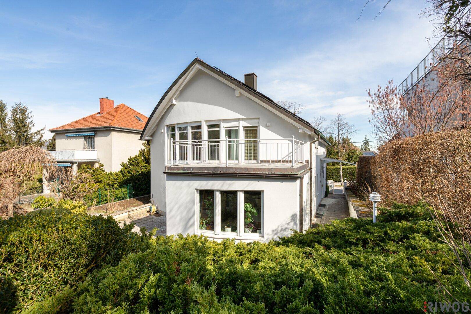 Seitenansicht des Hauses mit großen Fenstern, Balkon und gepflegter Heckenbepflanzung.