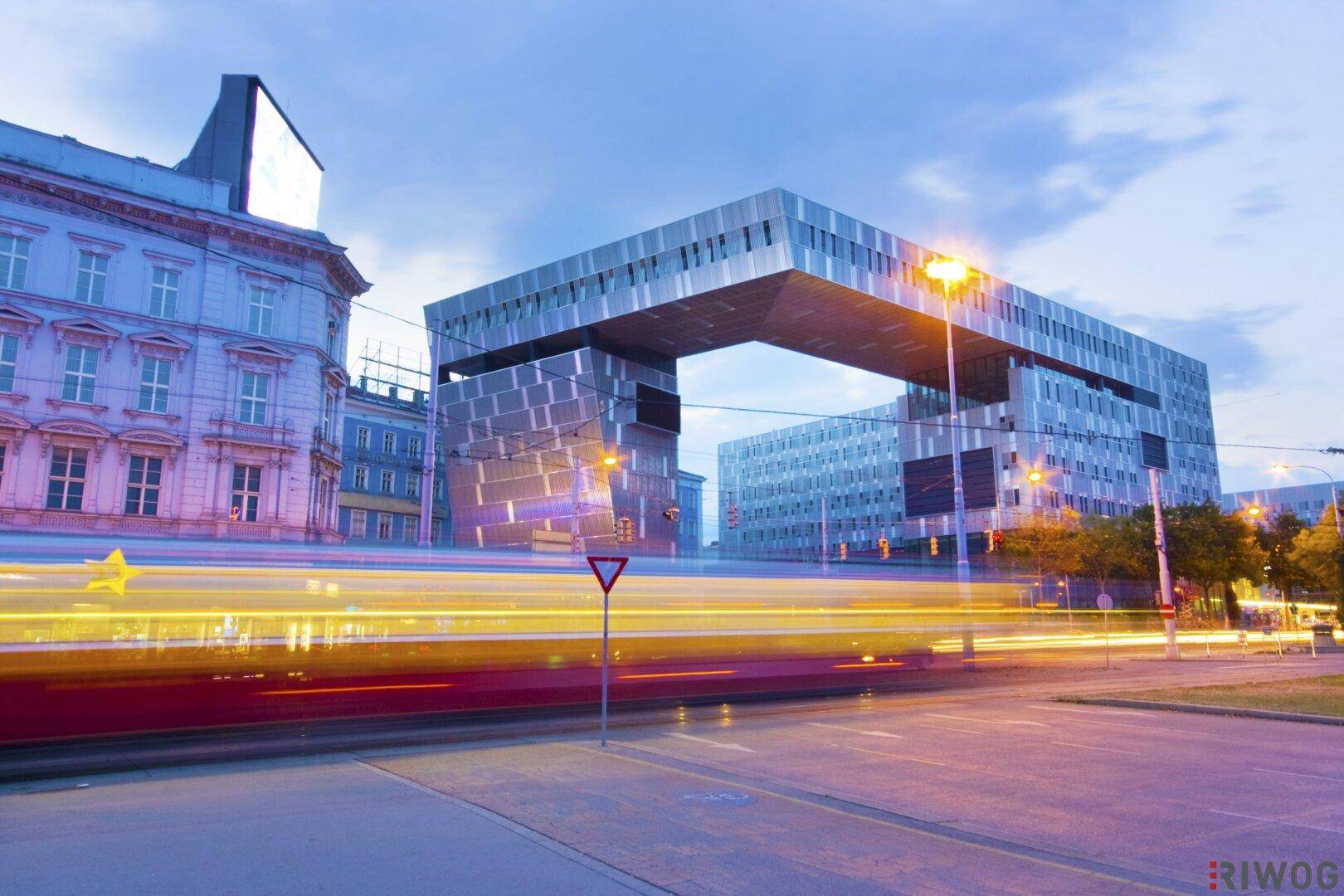 Moderne Architektur mit markanter Glasfassade und vorbeifahrendem Verkehr bei Dämmerung.