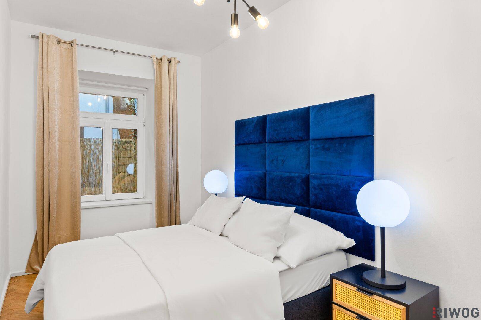 Helles Schlafzimmer mit Doppelbett, auffälligem blauen Kopfteil und Fenster mit beigen Vorhängen.