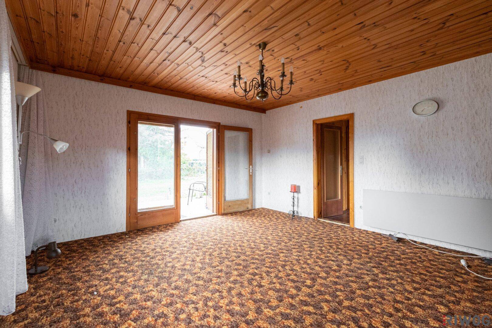 Geräumiges Wohnzimmer mit Holzdecke, gemustertem Teppichboden und Zugang zur Terrasse.