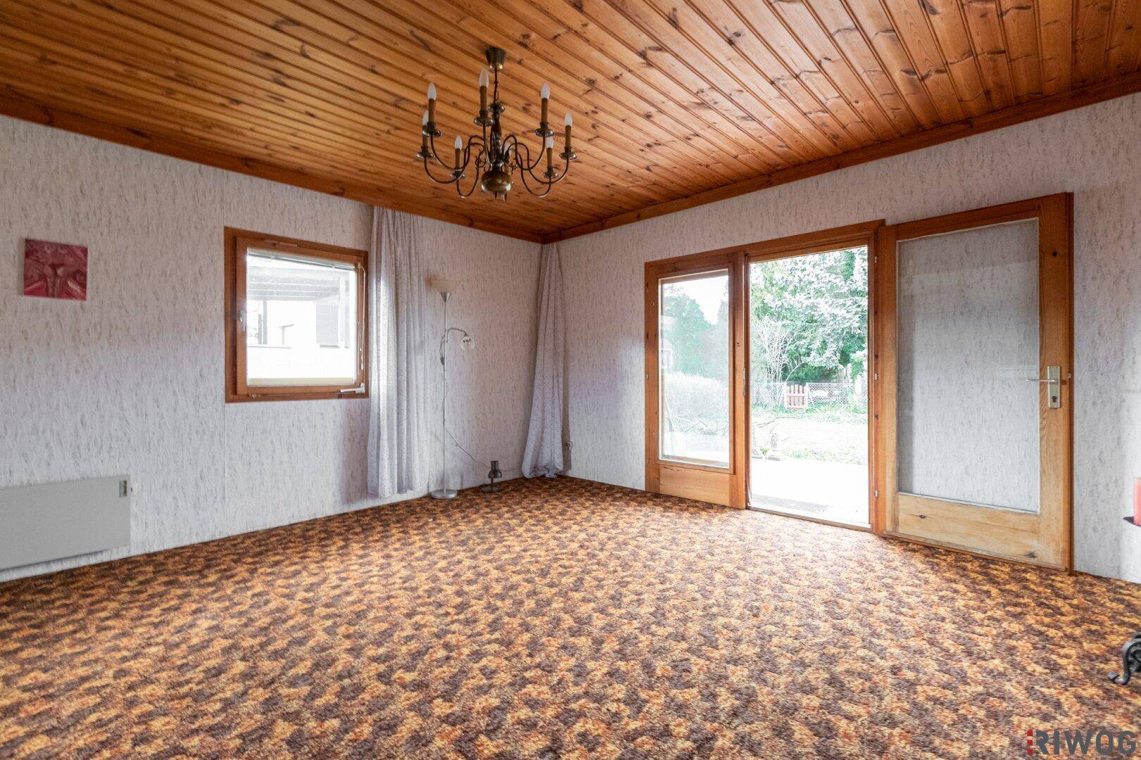 Helles Wohnzimmer mit Holzdecke, Teppichboden und großen Fenstern zum Garten.