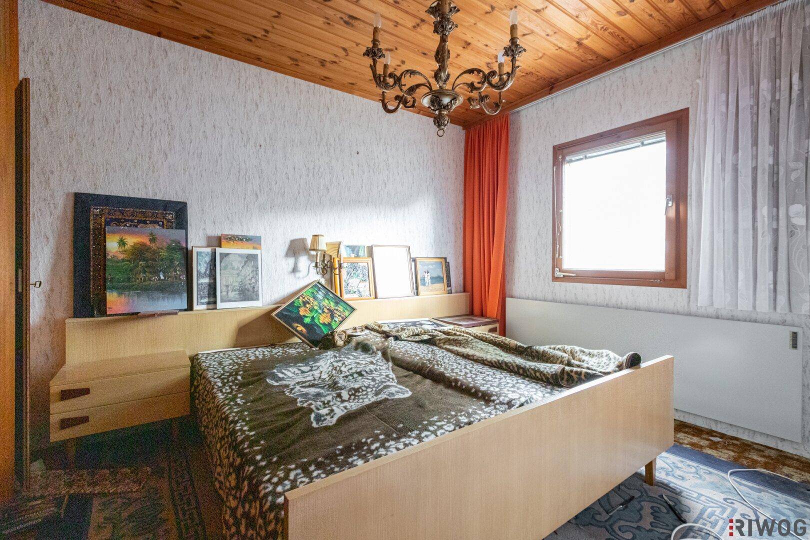 Schlafzimmer mit Holzbalkendecke, gemusterter Tapete und einem klassischen Kronleuchter.
