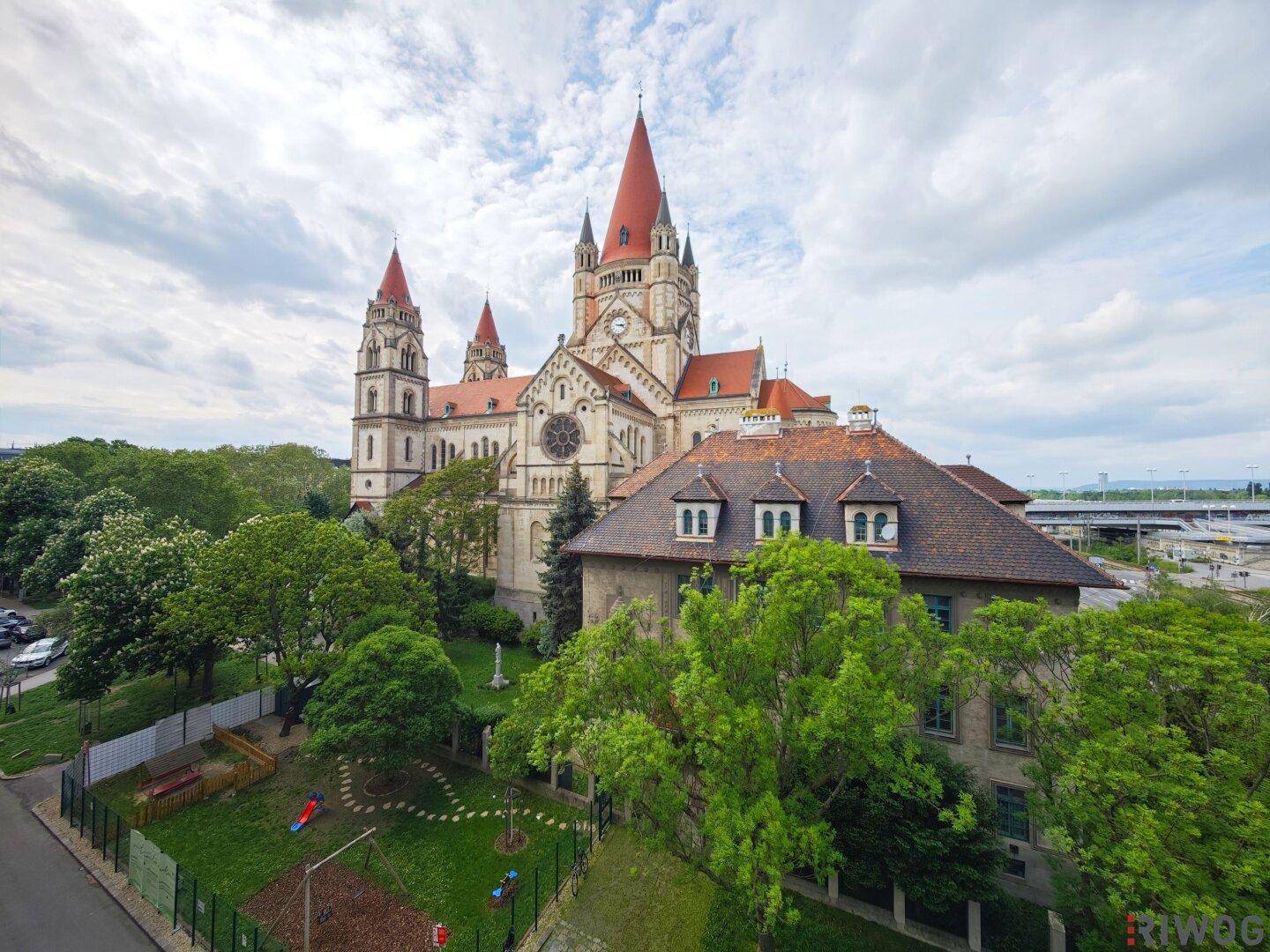 Panoramablick auf eine historische Kirche mit roten Dächern, umgeben von viel Grün und einem Spielplatz.