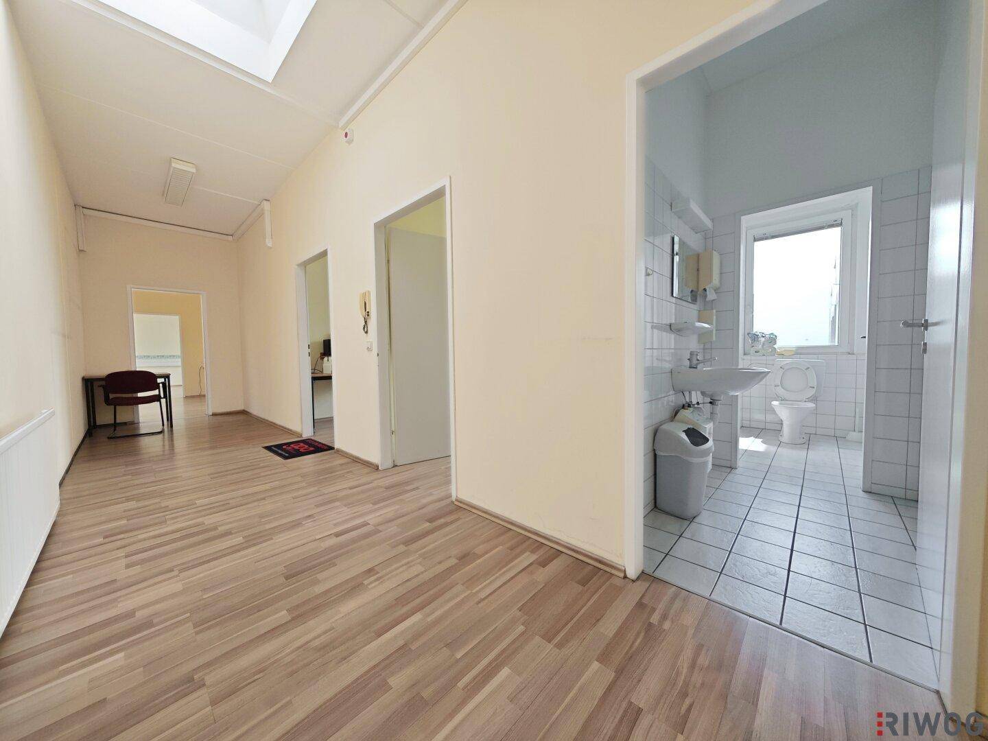 Flur mit Holzoptikboden, der zu einem gefliesten Badezimmer mit Fenster und weiteren Räumen führt.