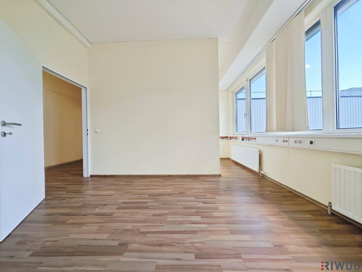 Helles Büro mit langen Fensterfronten, Holzoptikboden und Zugang zu weiteren Räumen im Gebäude.