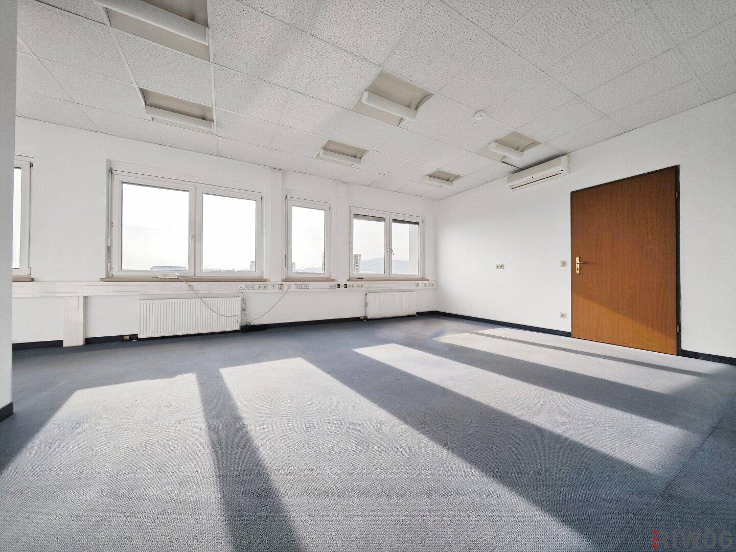 Leeres Büro mit blauem Teppichboden, Klimaanlage und natürlichem Lichteinfall durch die Fenster.