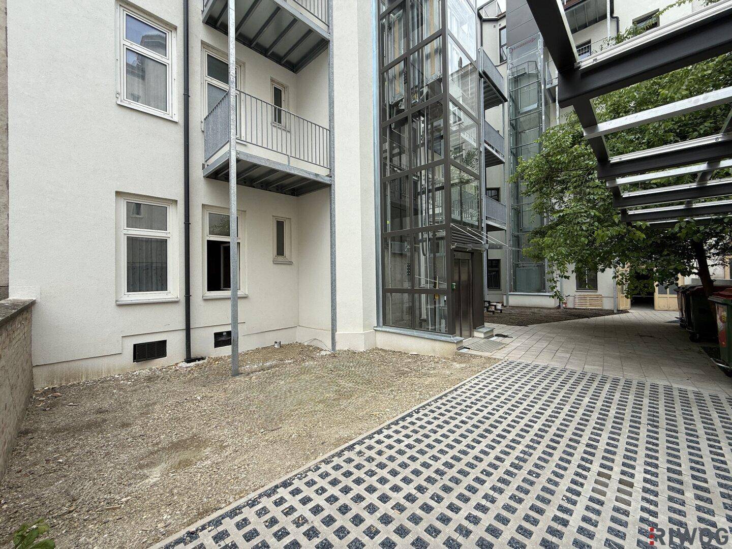 Innenhofbereich mit gepflastertem Weg, Kiesfläche und dem externen Glasaufzug des Gebäudes.