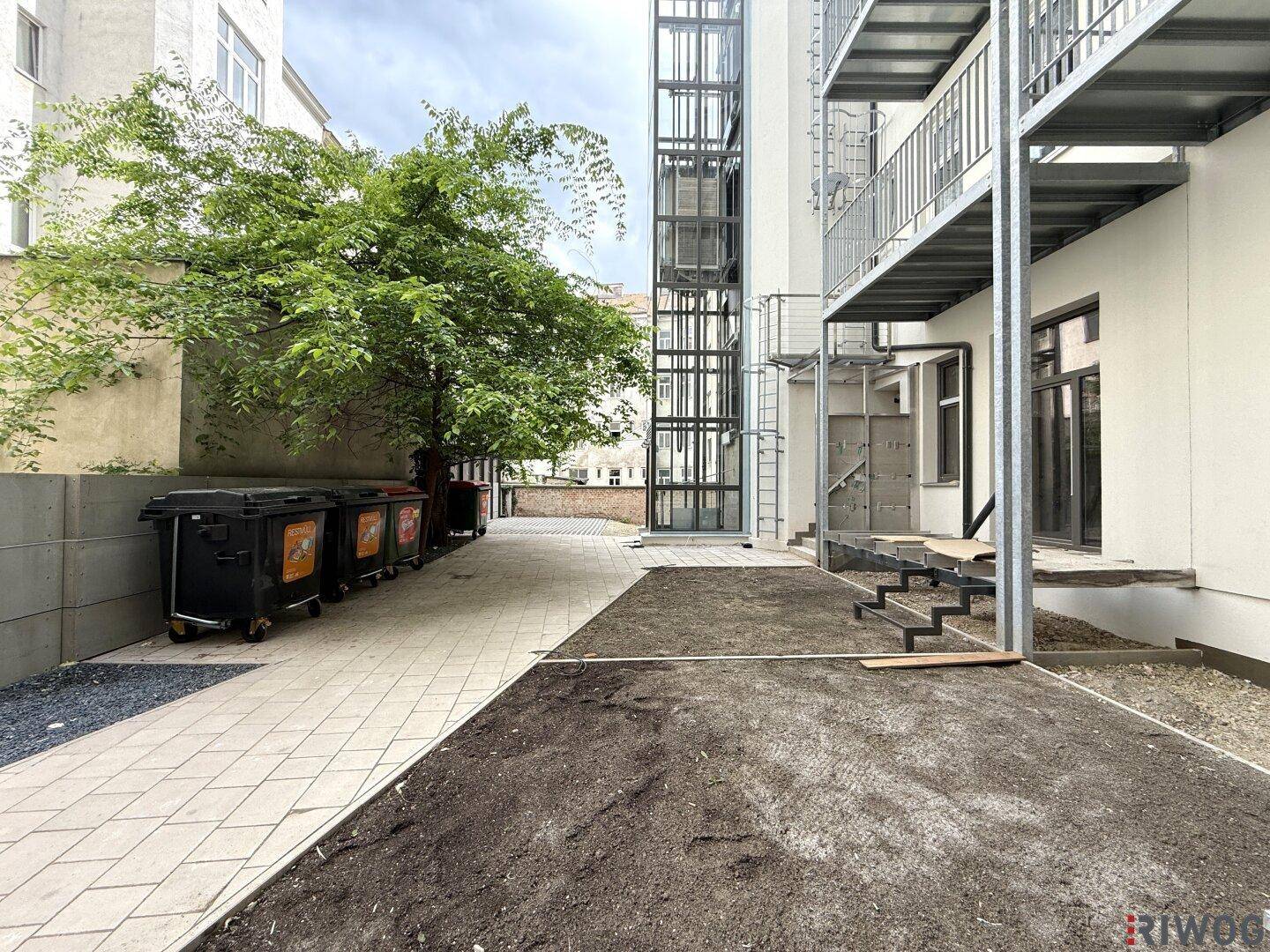 Außenansicht eines Gebäudes mit modernem Glasaufzug, Treppen und einem Baum im Vordergrund.