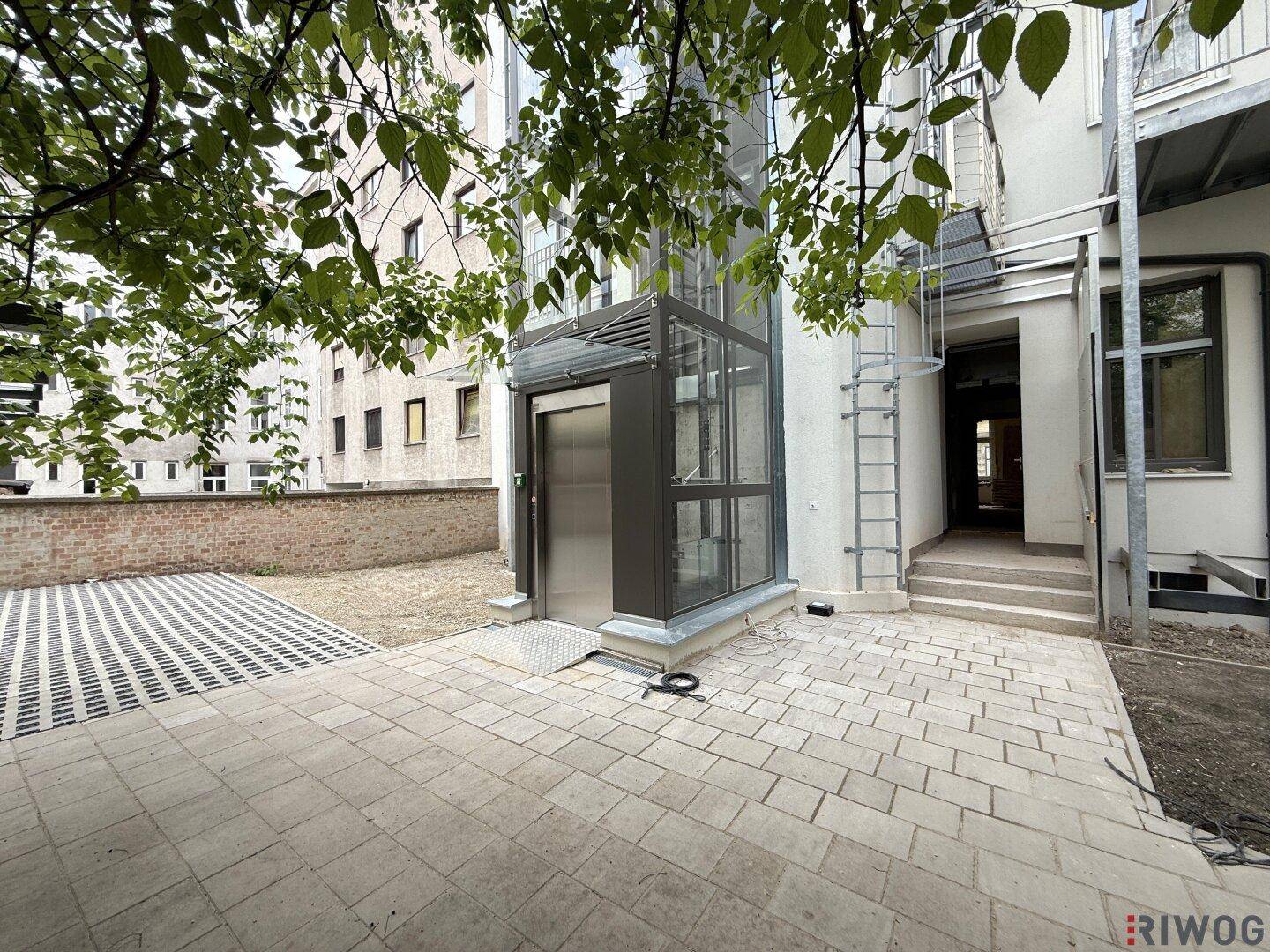 Außenbereich mit einem modernen Glasaufzug, gepflastertem Boden und Zugang zum Gebäude.