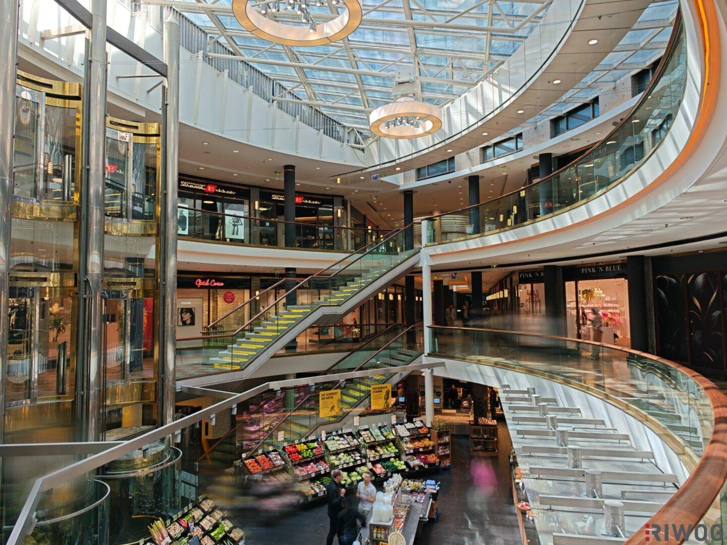 Modernes Einkaufszentrum mit Glasdach, Rolltreppen und Geschäften auf mehreren Ebenen.