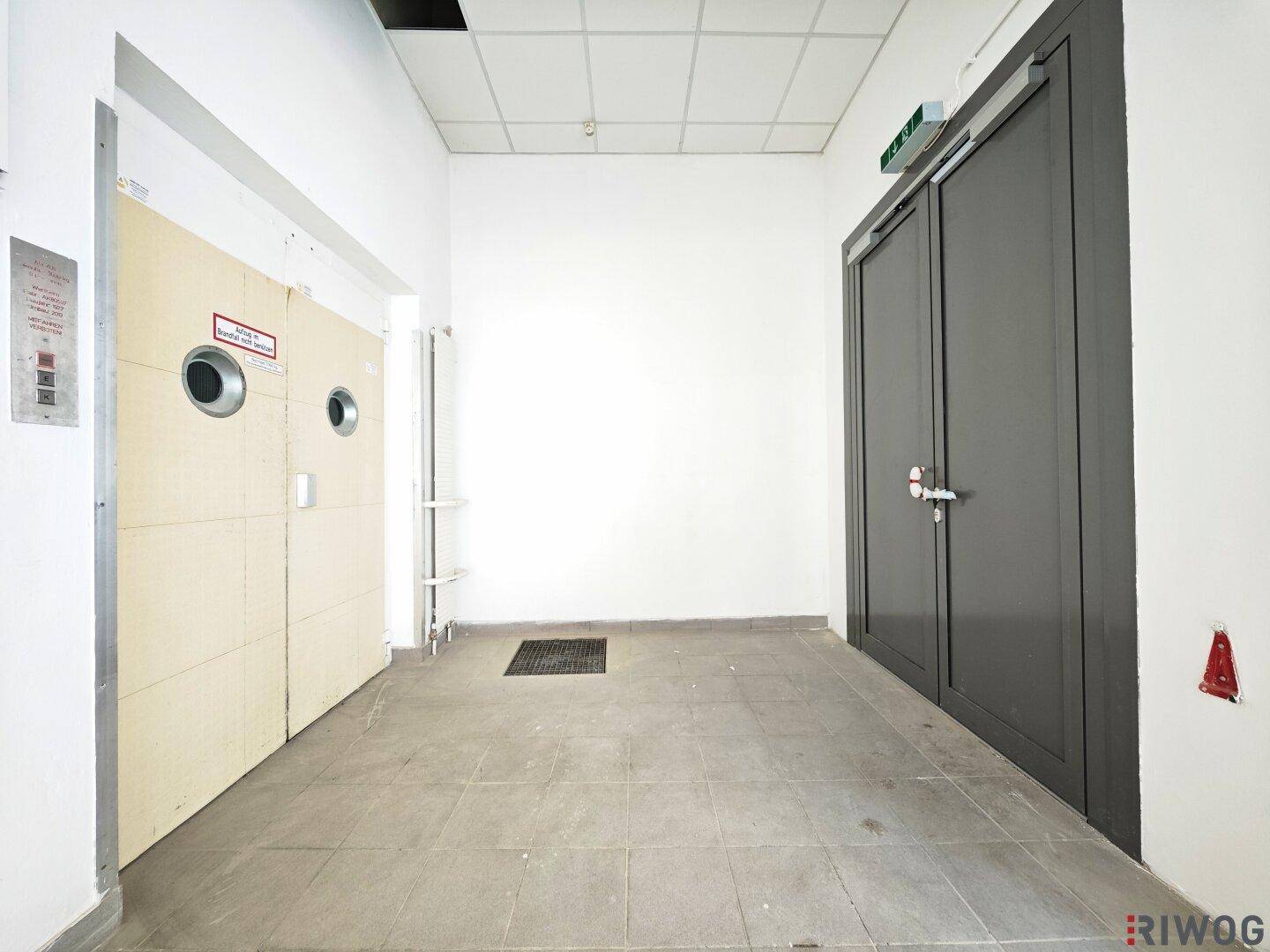 Flur mit Fliesenboden, zwei großen Türen und einem Aufzugsknopf an der Wand.