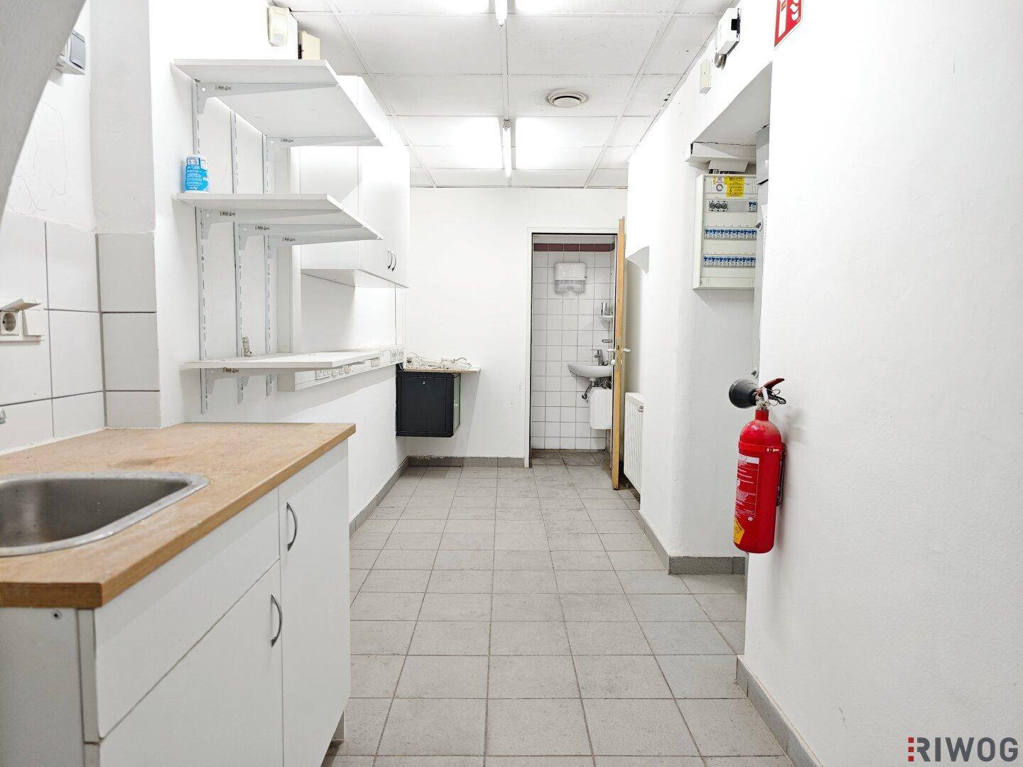 Kleine Teeküche mit Spüle, Arbeitsplatte und Regalen, angrenzend an eine Toilette.