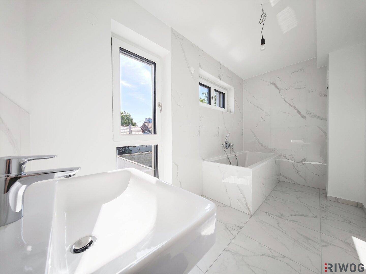 Modernes Badezimmer mit weißen Marmorfliesen, Badewanne und großem Fenster für Tageslicht.